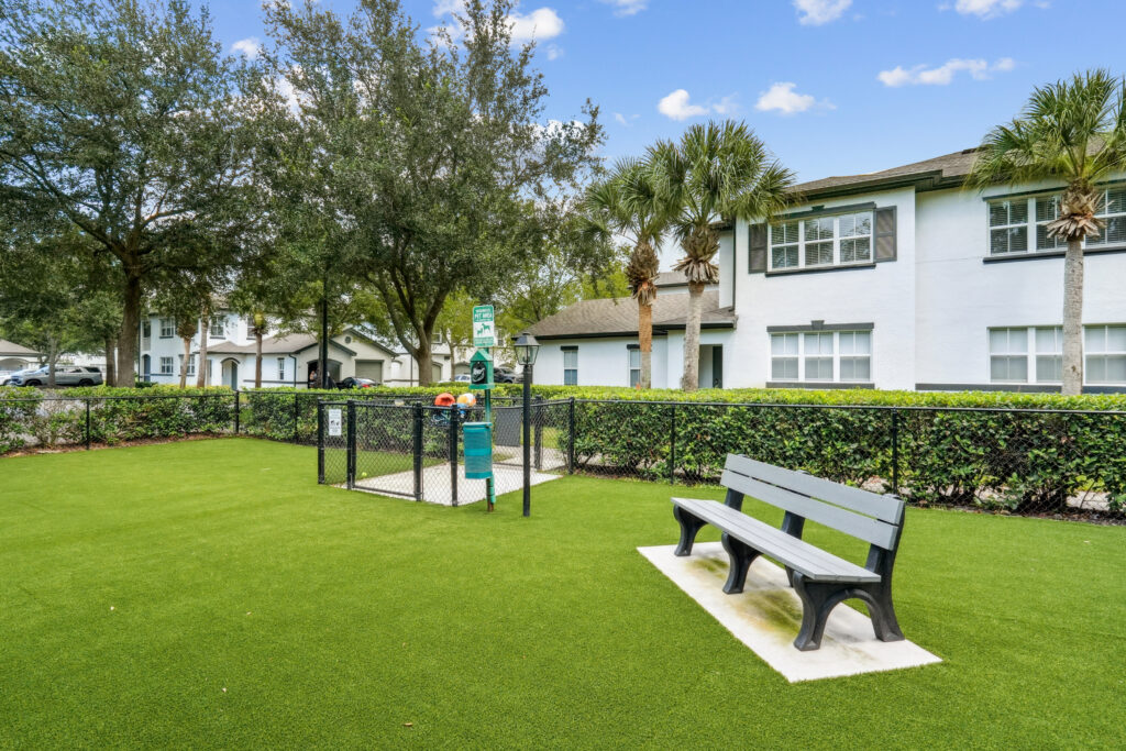 Lush grass, bench, and gated entrance to dog park in front of white buildings and trees