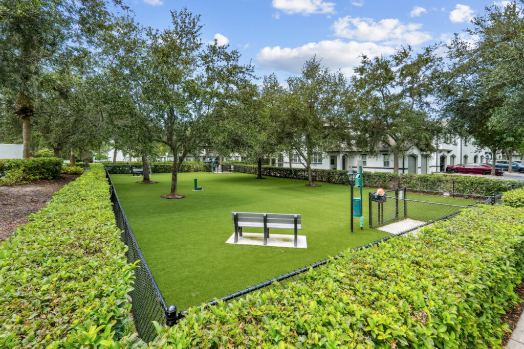 Dog park surrounded by hedges, with lush grass, bench, and gated entrance to park dotted with trees