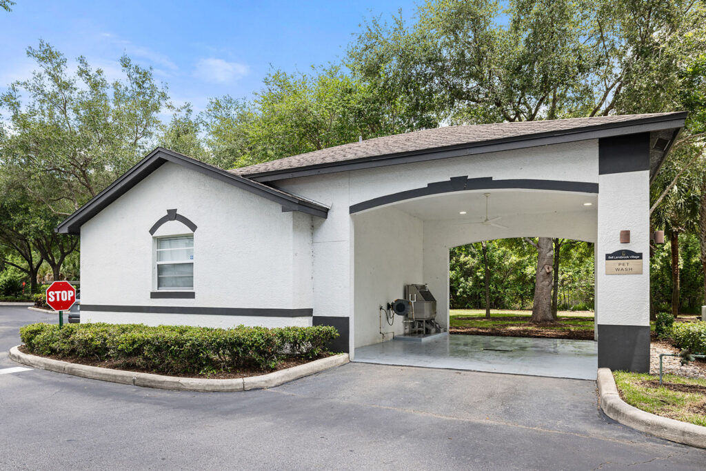 A small, white building with black trim features an open garage area containing cleaning equipment. It is surrounded by trees and greenery, with a stop sign visible on the left side of a paved road.