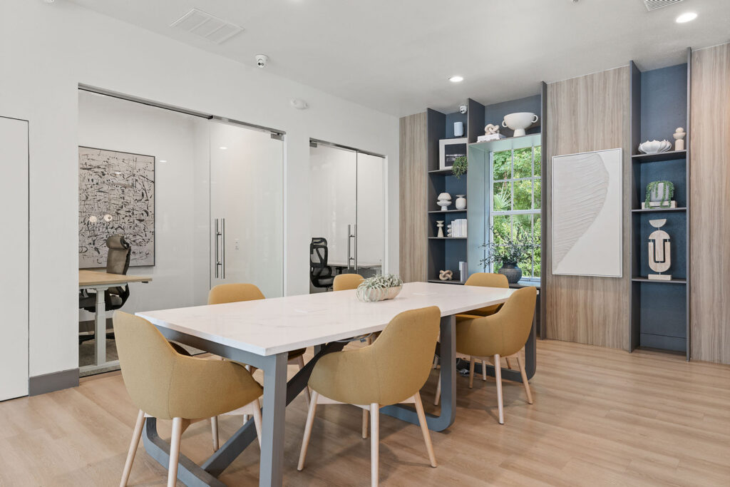 A modern meeting room with a white table, six mustard-yellow chairs, wooden flooring, built-in shelves with decor, and glass-walled offices in the background. Large windows let in natural light.