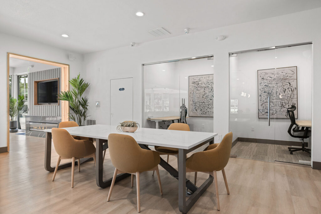 Modern conference room with a white table, five tan chairs, light wood flooring, potted plants, and abstract art on the walls. Adjacent glass-enclosed office spaces and a lounge area with a TV are visible.