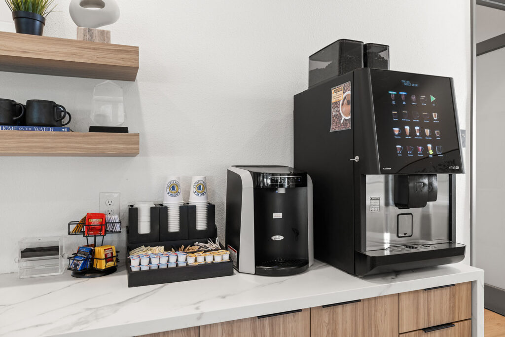 A countertop coffee station with a large coffee machine, a smaller brewer, disposable cups, creamers, sugar packets, stir sticks, and shelves with mugs and supplies against a white wall.