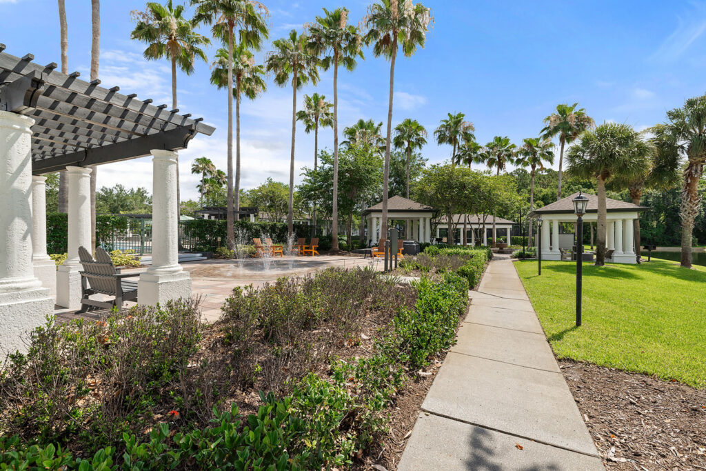 A paved walkway runs through a landscaped garden with bushes and palm trees, leading to white gazebos and a shaded seating area under a pergola on a sunny day.