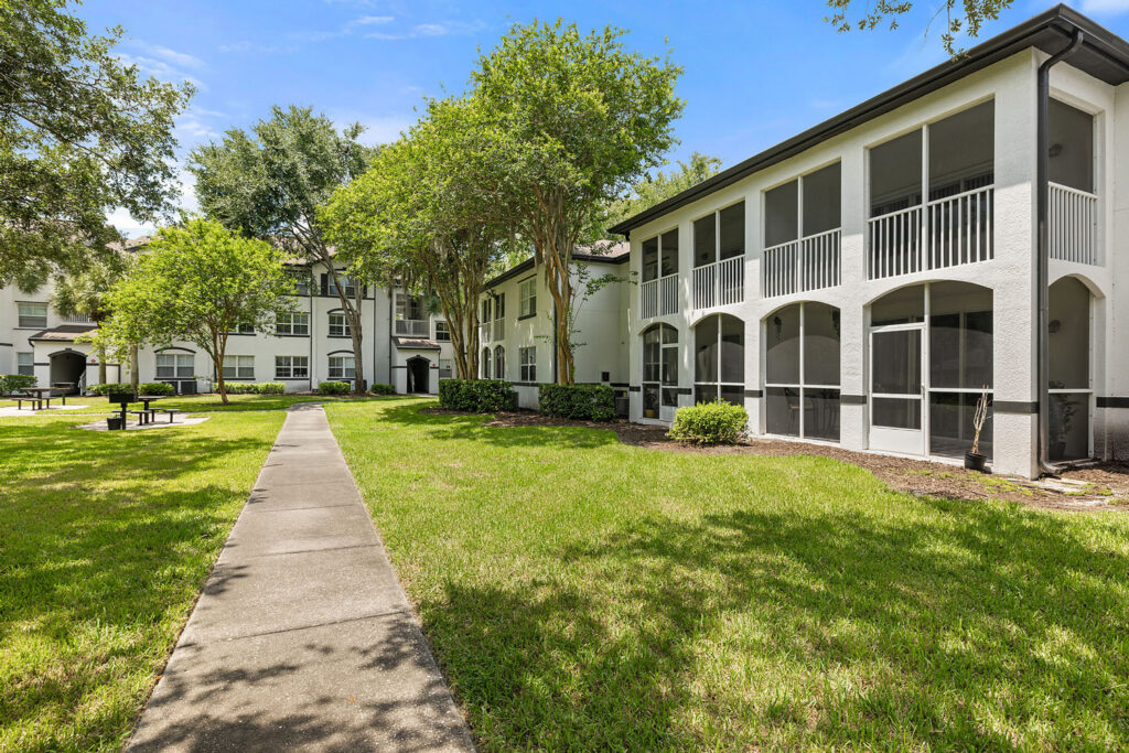 A walkway leads through a grassy courtyard with trees and picnic tables, bordered by white two-story apartment buildings with screened balconies on a sunny day.