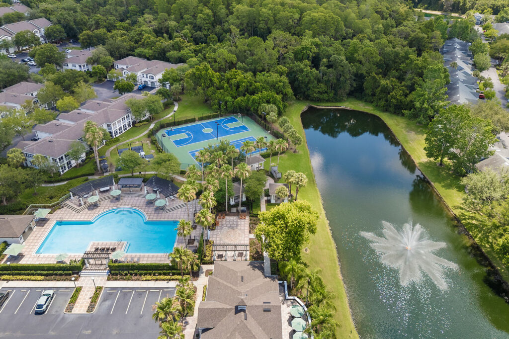 Aerial view of an apartment complex with a large swimming pool, sports courts, a pond with a fountain, parking area, and surrounding trees.