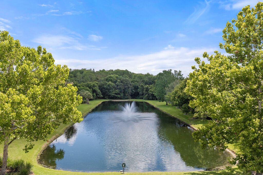 A small, serene pond with a central water fountain is surrounded by lush green grass and trees under a bright blue sky with wispy clouds.