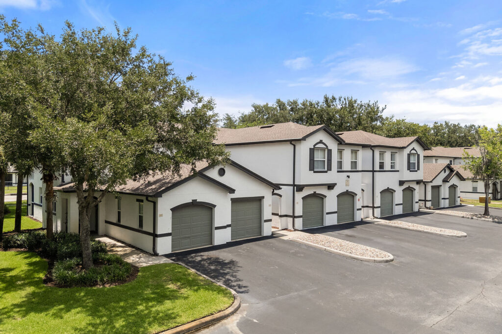 A row of modern, white townhouses with attached garages, black trim, and surrounded by green lawns and trees under a bright, blue sky. The parking area in front is empty.