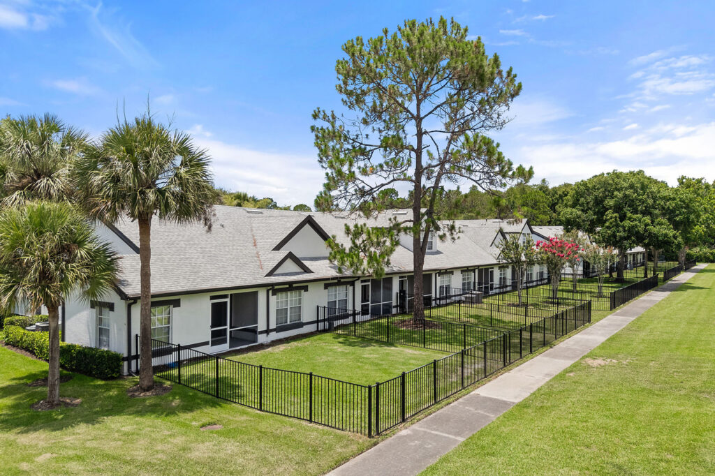A row of single-story, white residential buildings with gray roofs and black trim, each with fenced-in grassy yards, palm trees, and a sidewalk running alongside the property under a blue sky.