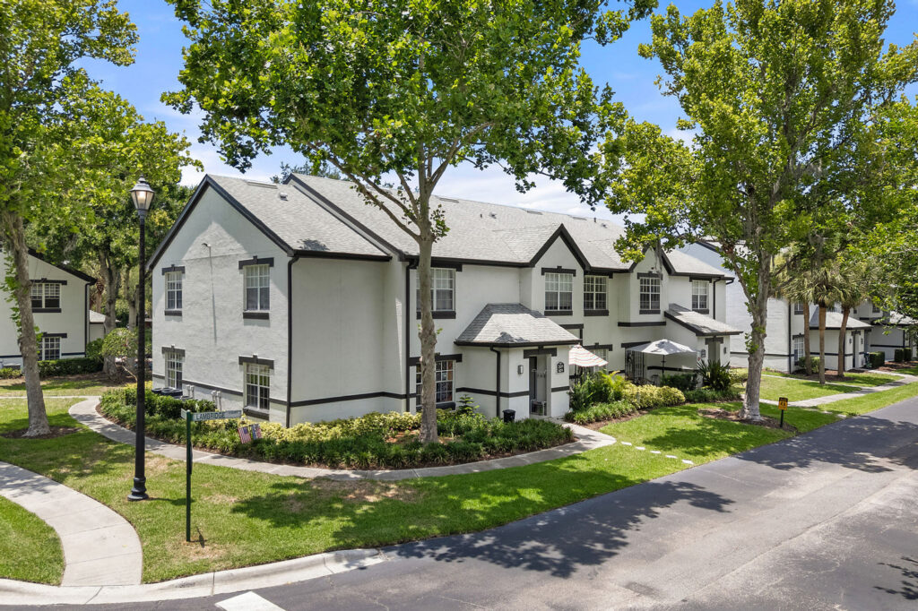 A two-story white townhouse with black trim, surrounded by green trees and landscaped bushes, sits on a curved street with sidewalks and street lamps on a sunny day.