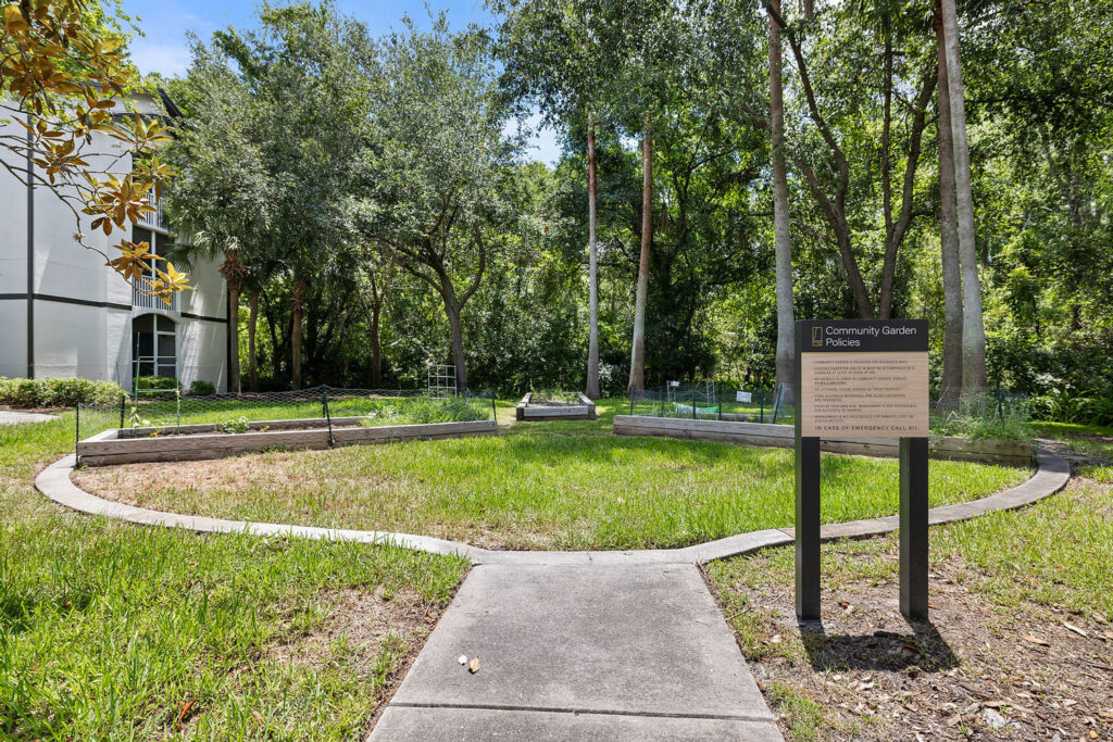A circular community garden with raised beds surrounded by trees, next to a sidewalk. A sign with garden policies stands at the entrance. The area is lush and green, bordered by a white building on the left.