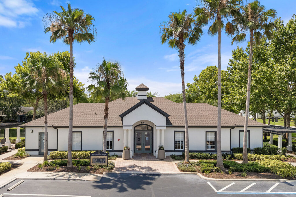 A single-story white building with brown roof sits behind a row of tall palm trees, surrounded by landscaped greenery and a parking lot under a bright blue sky.