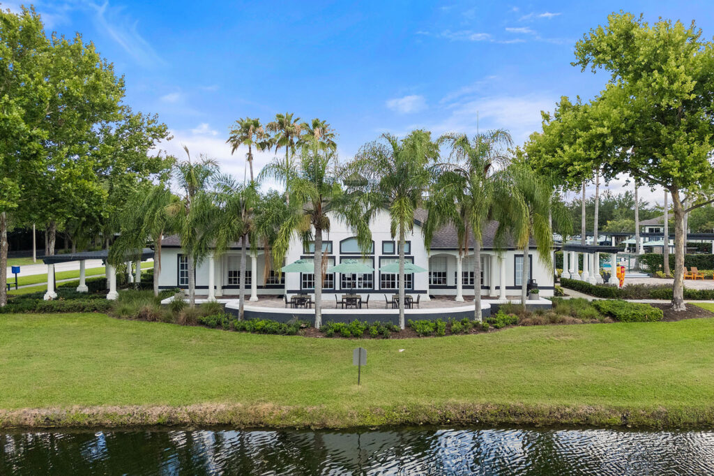 A white building with large windows and a shaded patio area, surrounded by palm trees and greenery, sits beside a calm pond under a bright blue sky.
