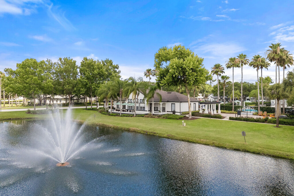 A large water fountain sprays in the middle of a pond near a manicured lawn, with a white building, palm trees, and a swimming pool in the background under a bright blue sky.