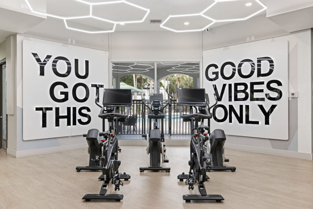 Three stationary exercise bikes face mirrors in a bright, modern gym with wood floors. Large signs on the walls read YOU GOT THIS and GOOD VIBES ONLY. Geometric ceiling lights hang above.