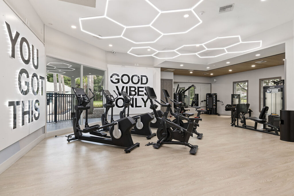 Modern gym with cardio and strength equipment, light wood floors, large windows, and motivational signs on the walls reading YOU GOT THIS and GOOD VIBES ONLY. Ceiling features geometric LED lighting.