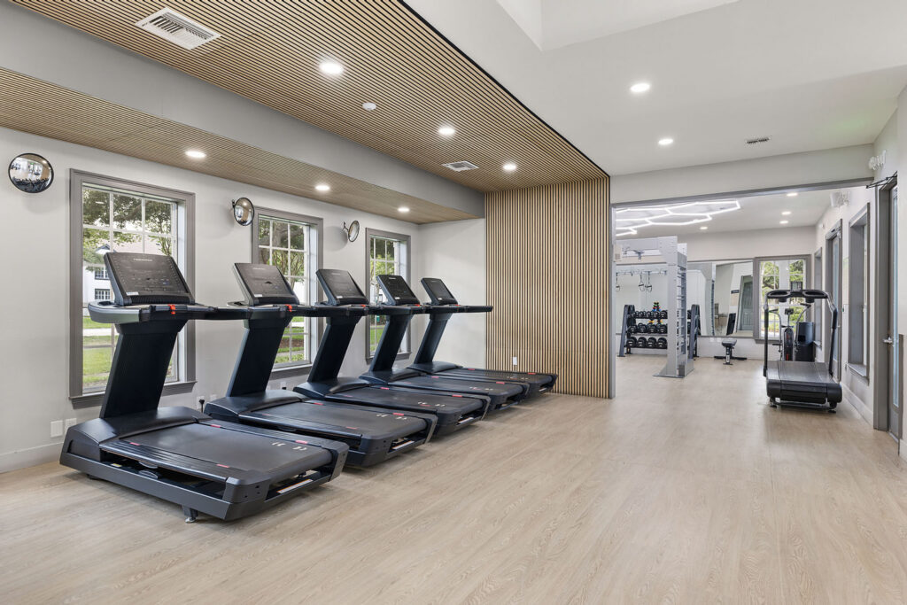 Modern gym with four treadmills lined up near large windows, light wood flooring, a wooden accent wall, and strength training equipment visible in the background. The room is brightly lit with overhead lights.
