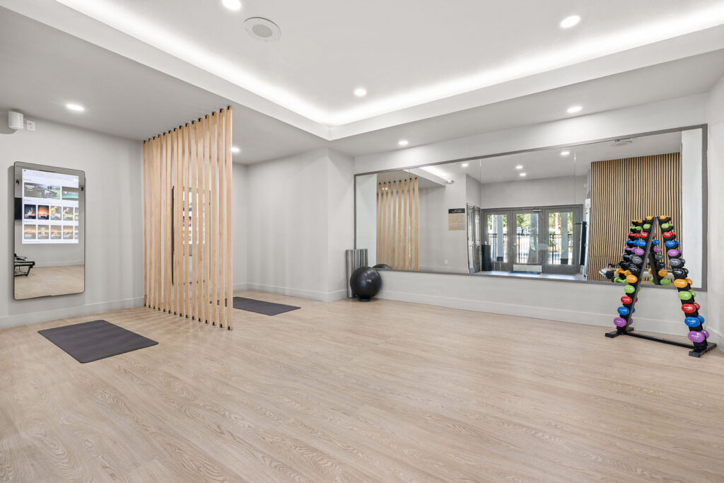 A bright, modern fitness room with light wood floors, floor-to-ceiling mirrors, yoga mats, a stability ball, foam rollers, and a rack of colorful dumbbells against the wall. Vertical wood slats act as a partition.