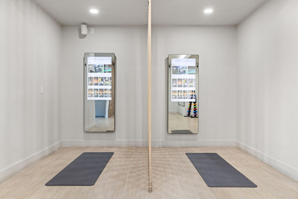 A modern fitness studio with two yoga mats placed on light wood floors, a central pole, and two full-length mirrors on the wall. Exercise equipment and posters are visible through the mirrors reflection.