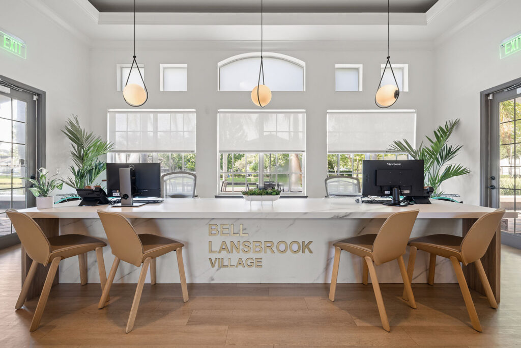 Modern office reception area with a marble desk, four tan chairs, two computers, decorative plants, large windows, and pendant lights. BELL LANSBROOK VILLAGE is displayed on the front of the desk.