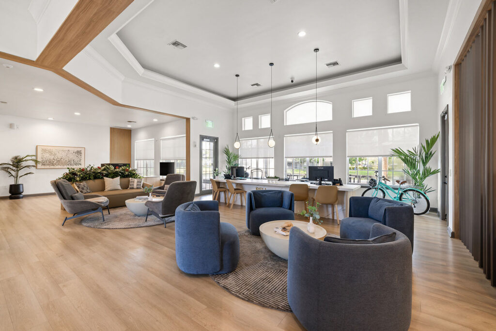 Modern, spacious lounge area with blue armchairs around white tables, large windows letting in natural light, indoor plants, a bicycle, and wooden flooring with contemporary decor.