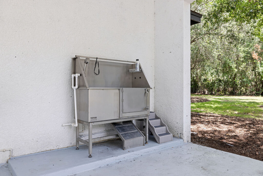 A stainless steel dog washing station with steps is positioned against a white exterior wall, adjacent to a grassy yard with trees in the background.