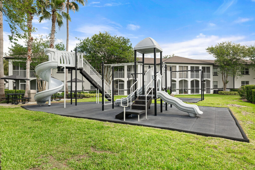 A modern playground with a spiral slide, straight slide, and climbing stairs sits on a rubber mat, surrounded by grass and trees, with apartment buildings in the background.