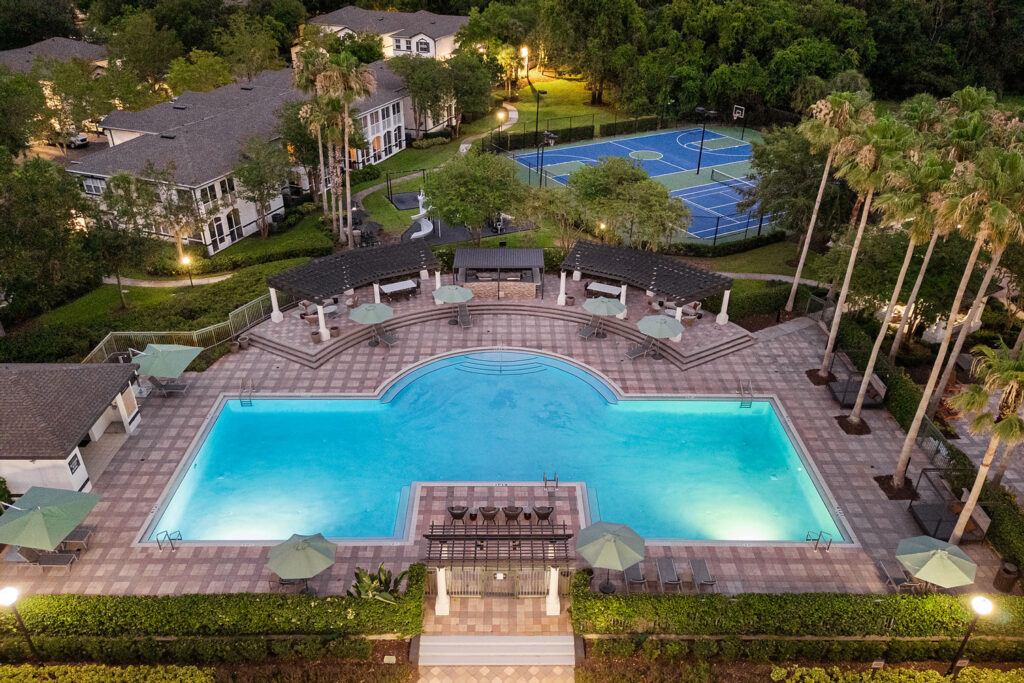Aerial view of a large, illuminated outdoor swimming pool with lounge chairs and umbrellas, surrounded by palm trees, landscaped gardens, and nearby tennis courts and residential buildings.