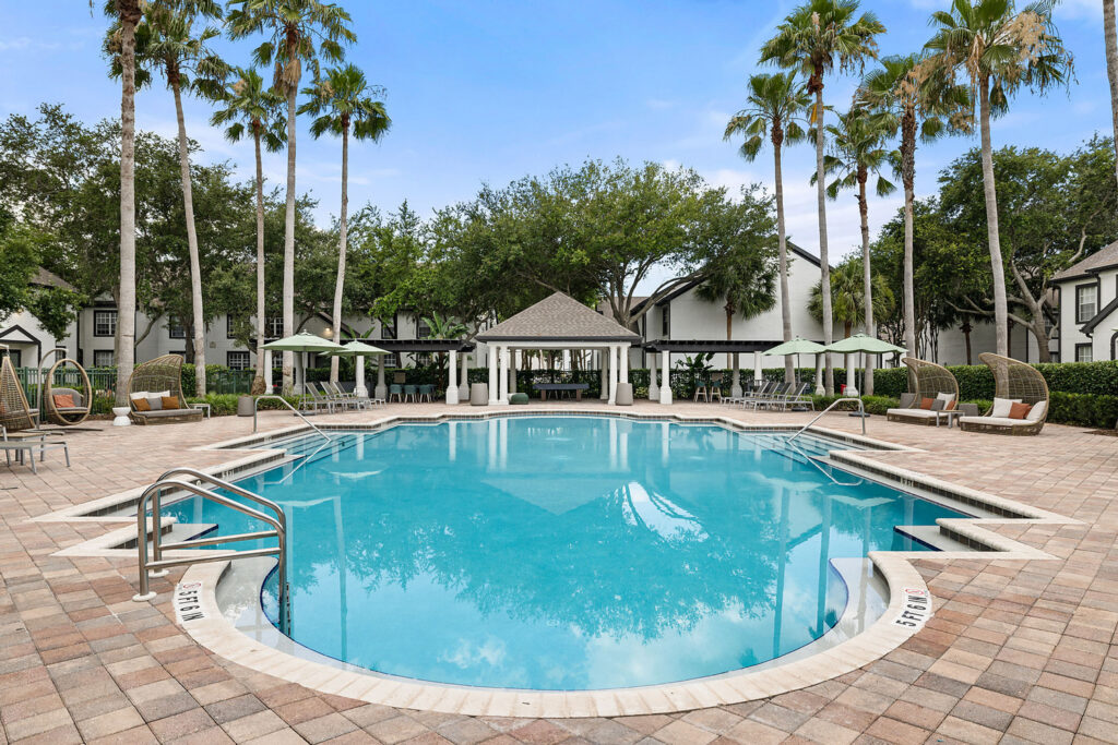 A large outdoor swimming pool surrounded by lounge chairs, cabanas, palm trees, and brick paving. White residential buildings and greenery are visible in the background under a blue sky.