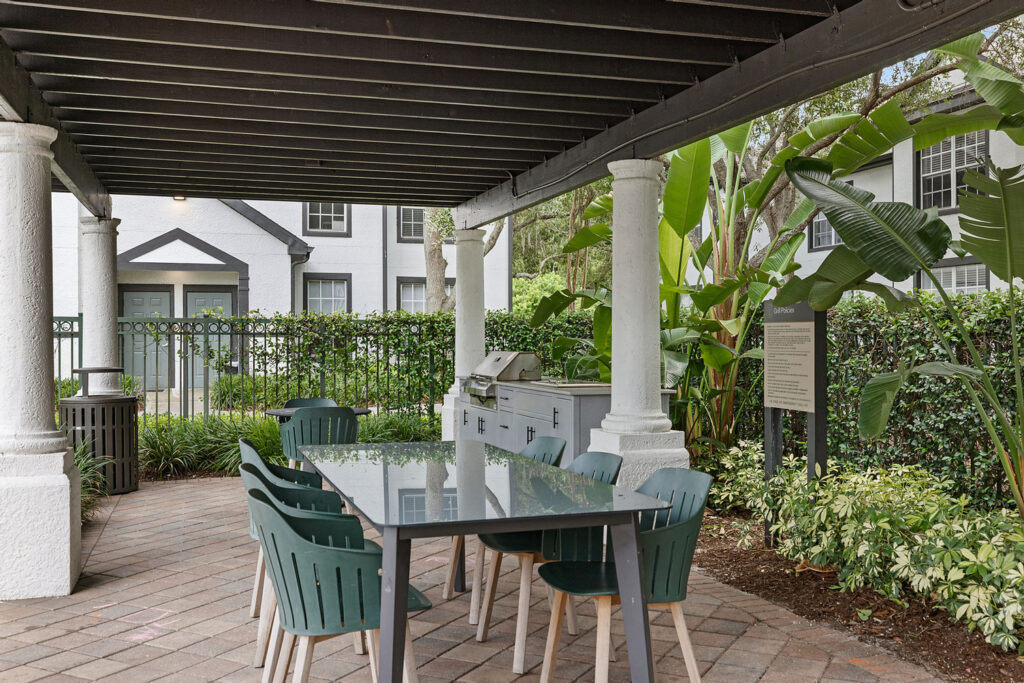 Outdoor patio area with a long table, green chairs, a grill, and a shaded pergola. Lush greenery and tropical plants surround the space, with a white building visible in the background.