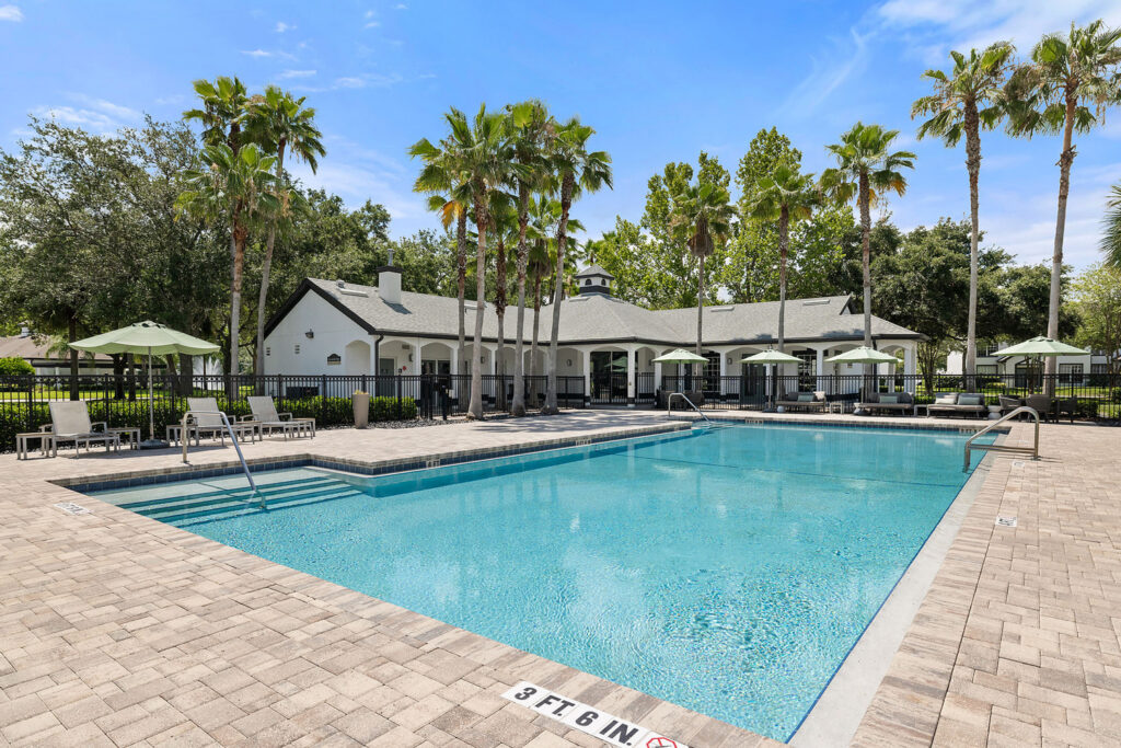Outdoor swimming pool surrounded by lounge chairs, green umbrellas, and tall palm trees, with a white clubhouse in the background under a blue sky. Pool deck is paved with light-colored bricks.