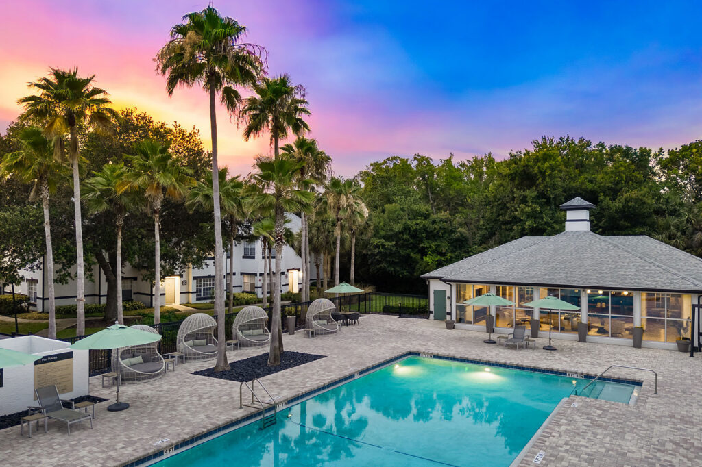 A swimming pool surrounded by lounge chairs, palm trees, and umbrellas, with a modern building and lush trees in the background under a colorful sunset sky.