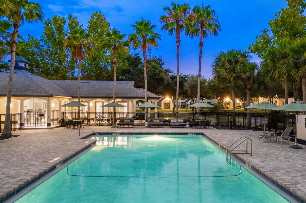 A swimming pool surrounded by lounge chairs and umbrellas, with palm trees and a clubhouse in the background, illuminated by evening lights. The sky is a deep blue, suggesting dusk or early evening.