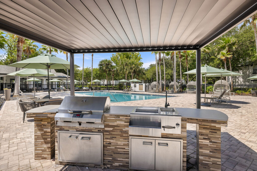 Outdoor kitchen with a stainless steel grill and sink under a covered patio, overlooking a swimming pool surrounded by lounge chairs, green umbrellas, and palm trees on a sunny day.