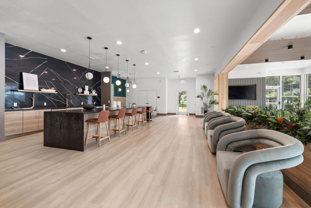 Modern communal lounge with a large kitchen island, wooden flooring, bar stools, a wall-mounted TV, lounge chairs, and abundant natural light from large windows. Decorative lighting and plants add to the contemporary decor.