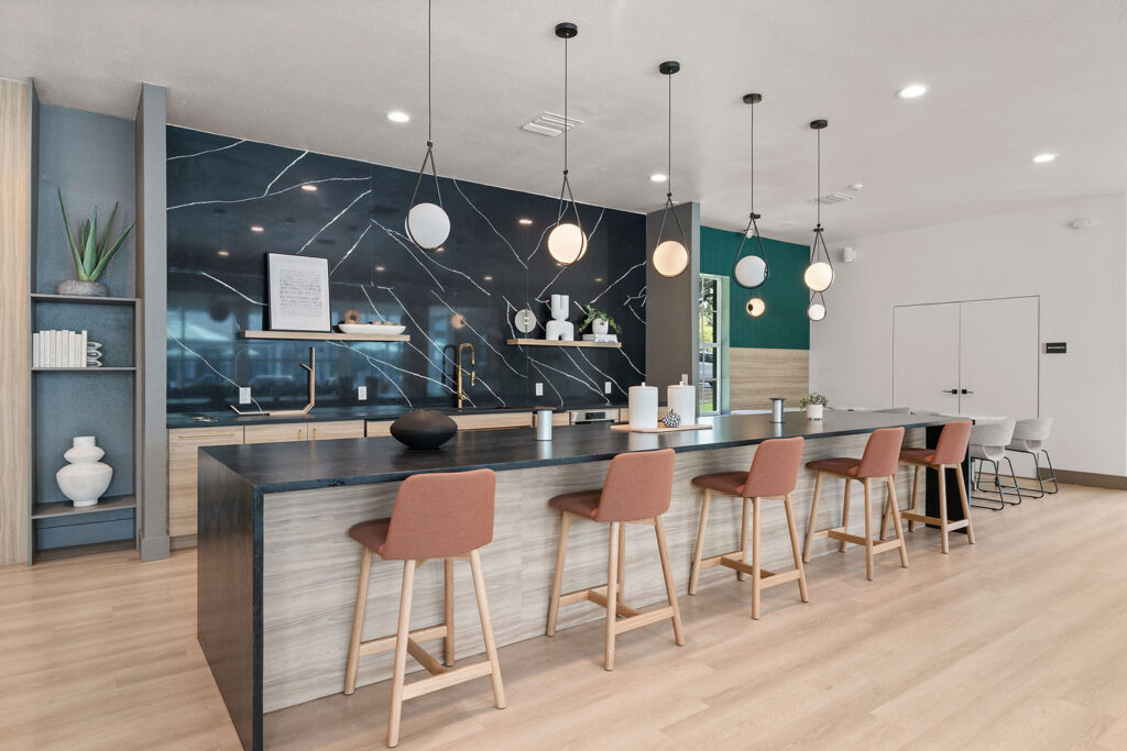 Modern kitchen with a long island, five pink bar stools, black marble backsplash, gold fixtures, pendant lights, decorative vases, and light wood floor. Shelves with books and plants are on the left side.