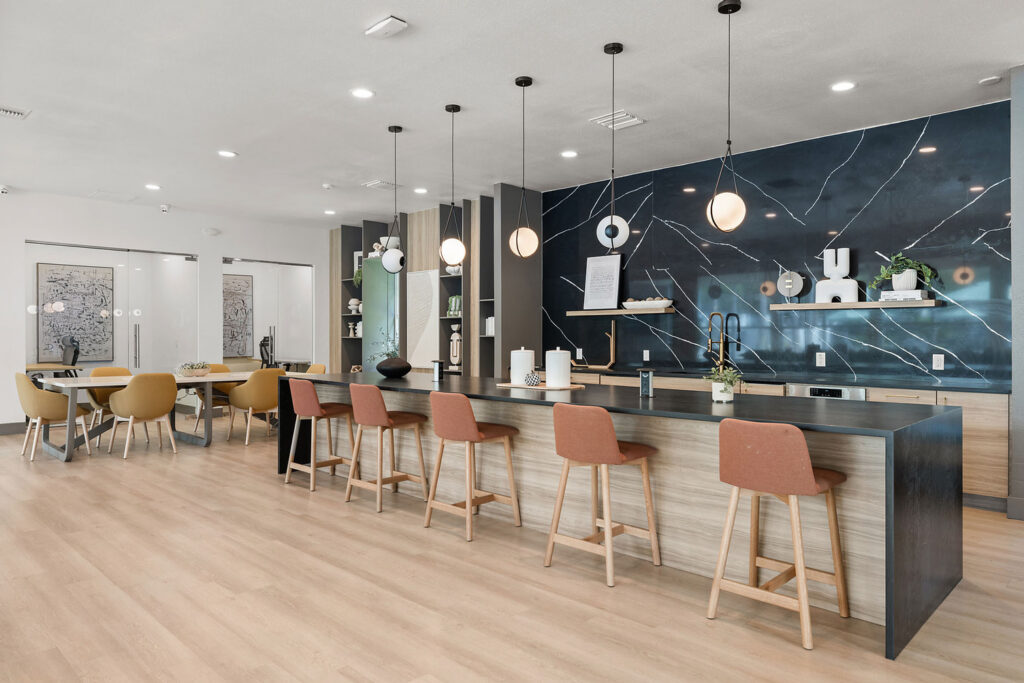 Modern communal kitchen with a large island, five brown barstools, pendant lights, and a dark marble-patterned backsplash. Dining tables with yellow chairs are visible in the background. Light wood flooring throughout.