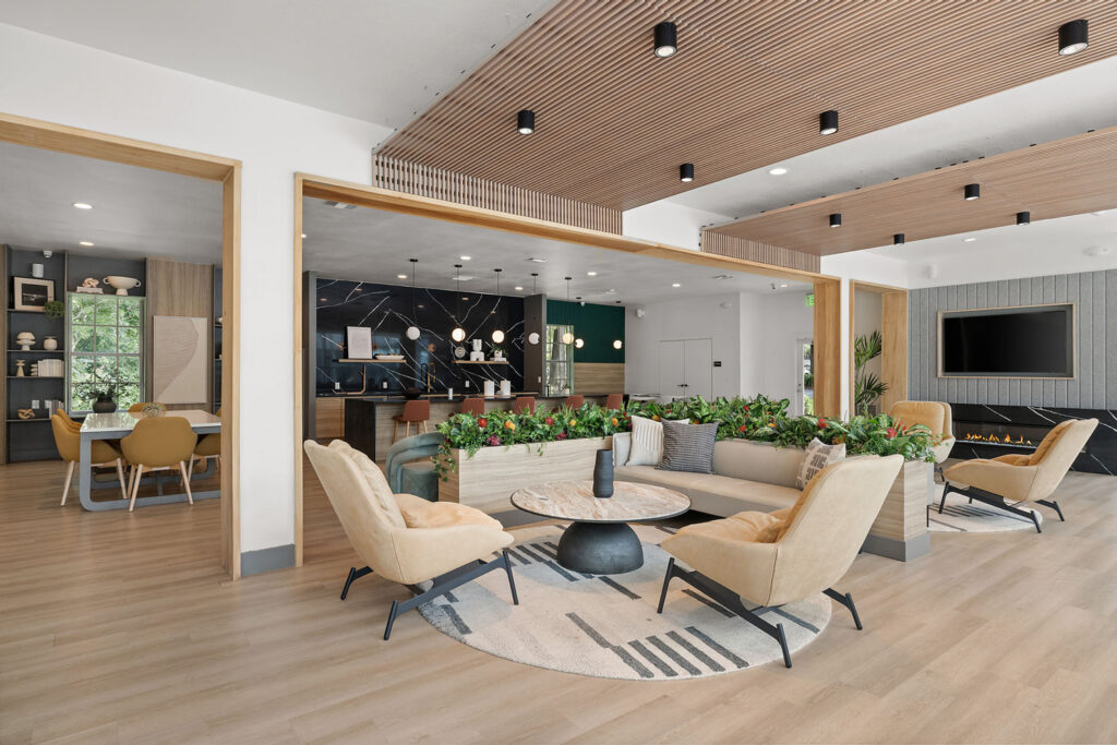 Modern lounge area with beige chairs, a round marble coffee table, an L-shaped sofa, plants, wood accents, and a fireplace; an open kitchen and dining area are visible in the background.