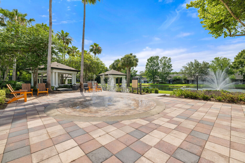 A circular splash pad with water jets surrounded by lounge chairs and trees, next to a pond with a fountain, under a bright blue sky.