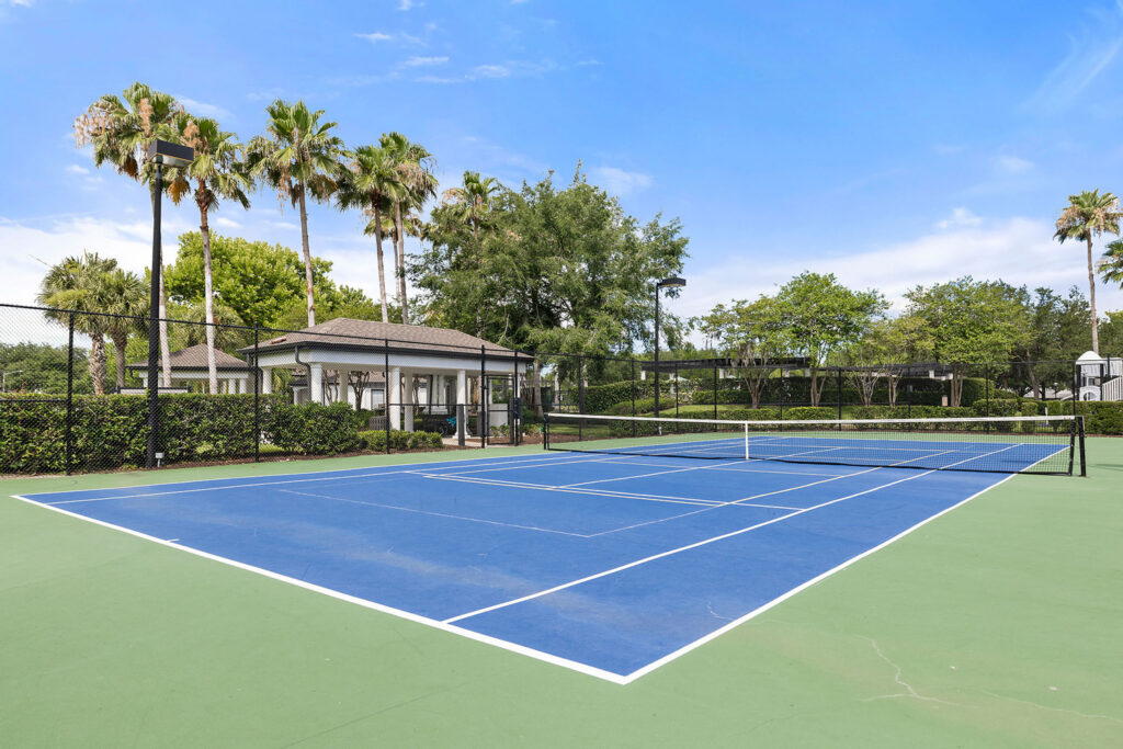 A bright blue tennis court with a green border stands empty outdoors, surrounded by a black fence, palm trees, and lush greenery under a clear blue sky.