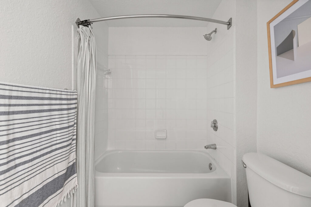 A clean, white bathroom with a bathtub and shower, white tile walls, a striped towel hanging on the left, a white toilet on the right, and a framed abstract art piece above the toilet.