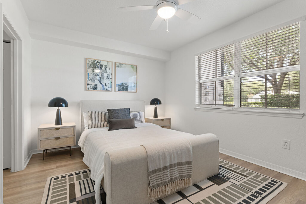 A bright bedroom with a bed, striped bedding, two bedside tables with black lamps, wall art above the bed, a ceiling fan, a rug underneath, and a large window with blinds letting in natural light.