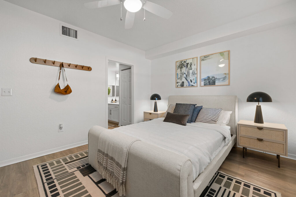 A modern bedroom with a beige upholstered bed, neutral bedding, and pillows. Two nightstands with lamps flank the bed. Art hangs above the headboard. A wall-mounted rack holds a bag. The room has a ceiling fan and patterned rugs.
