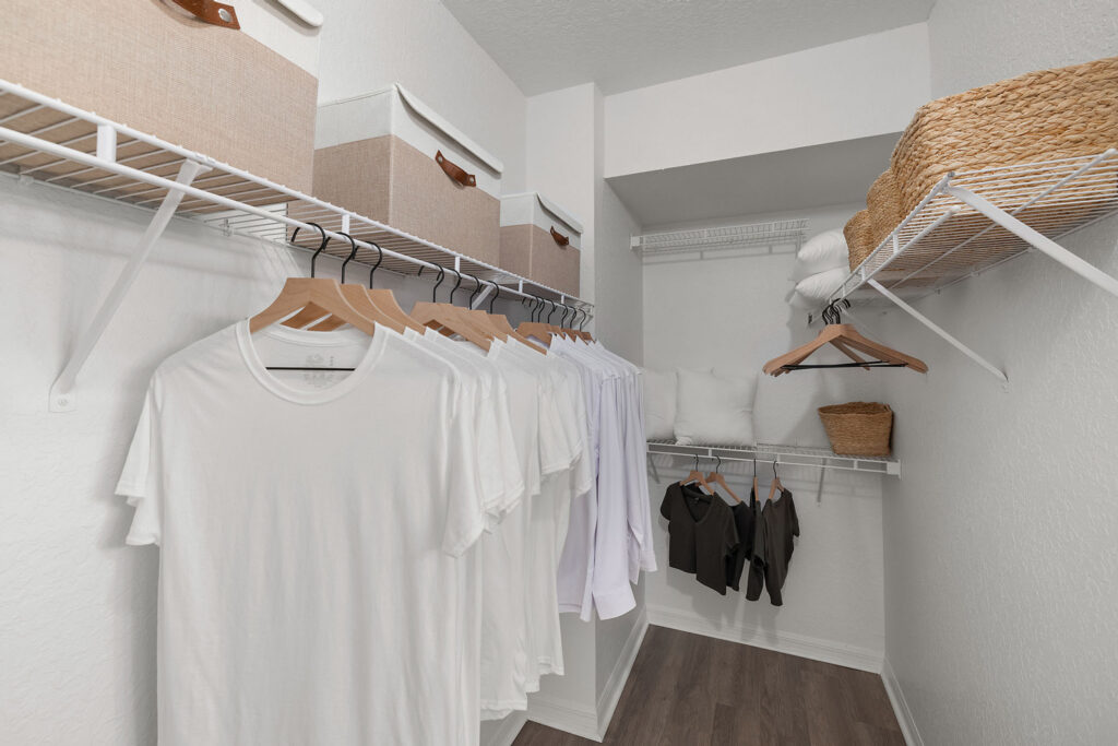 A neatly organized walk-in closet with white walls, light wood flooring, white shelving, several white shirts on wooden hangers, beige storage boxes, folded towels, pillows, and a woven basket on the shelves.