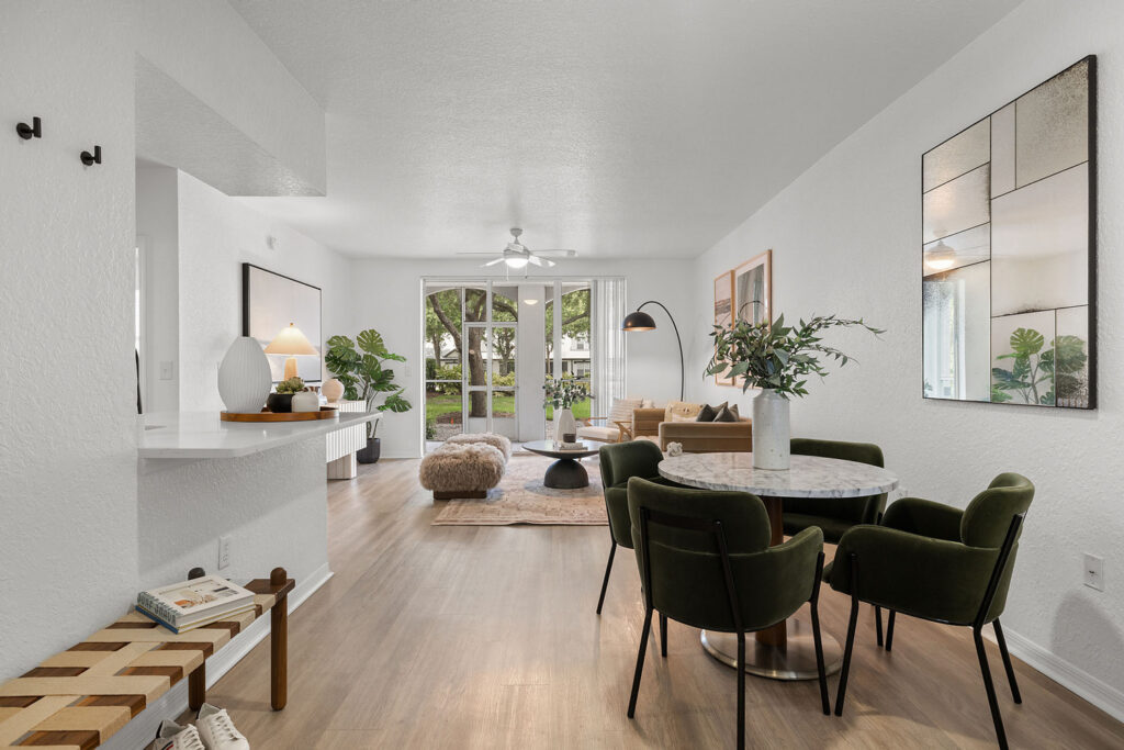 A modern, bright living and dining area with wooden floors, green velvet chairs around a marble table, a beige sofa, a large mirror, decorative plants, and glass doors opening to a patio with greenery outside.