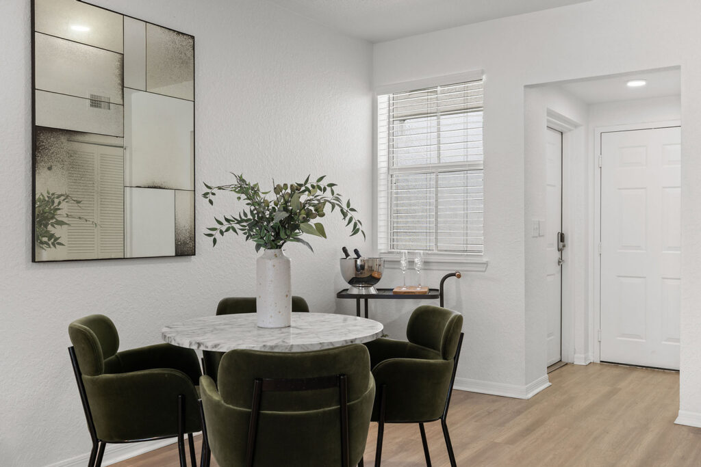 A modern dining area with a round marble table, four green velvet chairs, a large abstract wall mirror, a vase with greenery, a small bar cart, a window with blinds, and a white door on light wood flooring.
