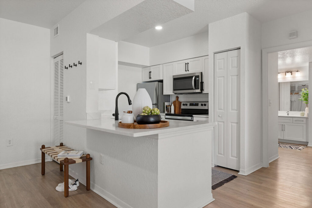 Modern kitchen with white cabinets, stainless steel appliances, and a breakfast bar with decorative items. A wooden bench sits by the wall, and a bathroom is visible through an open doorway in the background.