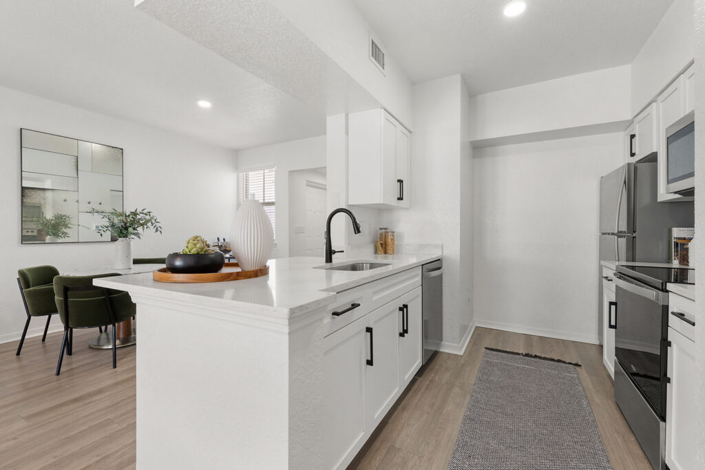 Modern kitchen with white cabinets, stainless steel appliances, and a black faucet. A tray with decorative items is on the counter. In the background, there is a dining area with green chairs and a large wall mirror.