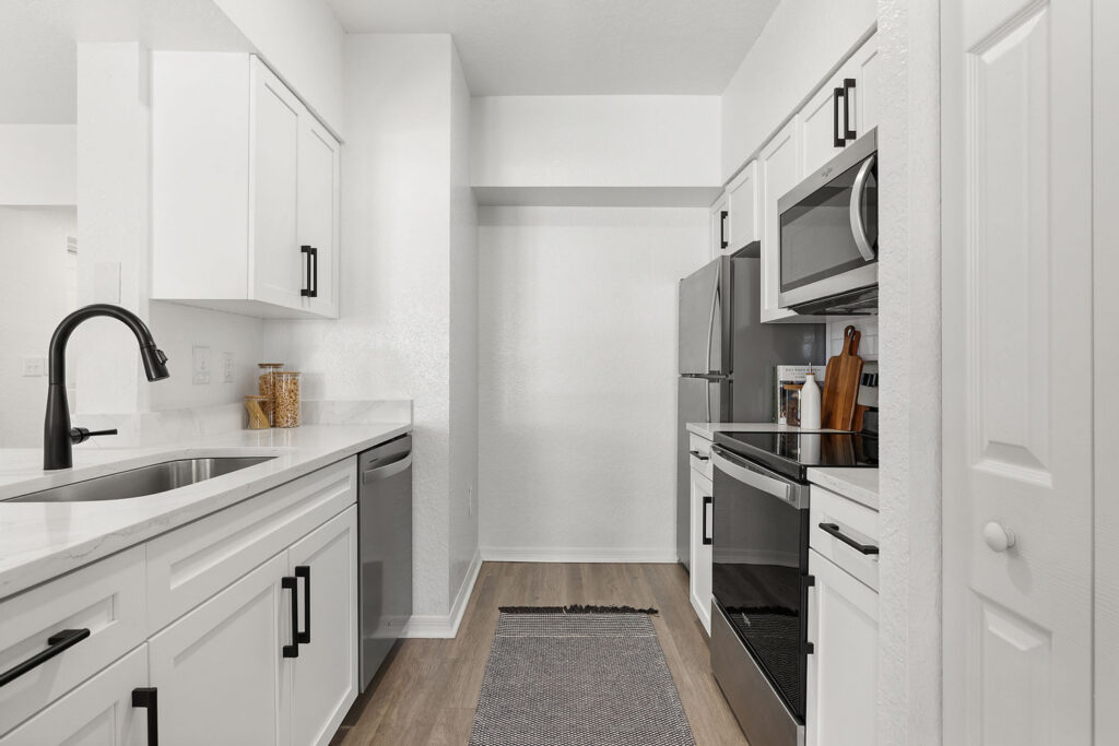 Modern, white galley kitchen with stainless steel appliances, black fixtures, marble countertops, white cabinets, a gray rug on wood flooring, and minimal decor including jars and cutting boards.