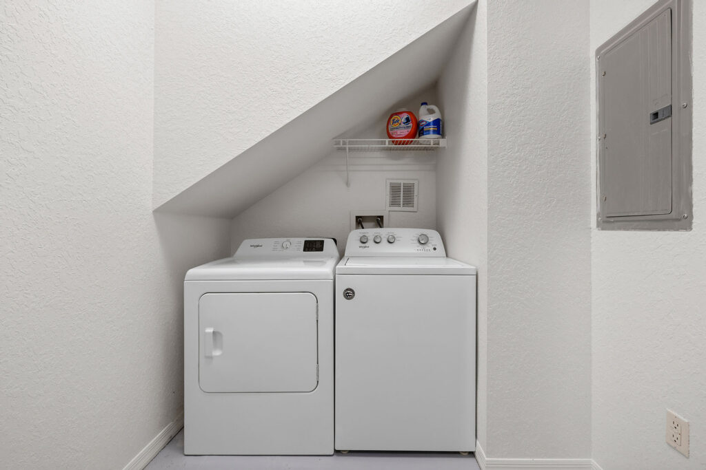 A small laundry area with a white washing machine and dryer under a sloped ceiling, a shelf above holding detergent and bleach, set against white textured walls.