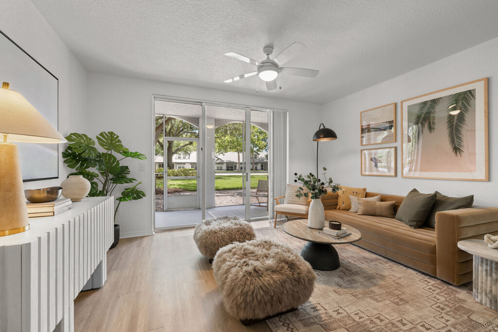 A modern living room with a tan leather sofa, furry ottomans, a round coffee table, large artwork, and sliding glass doors opening to a patio with greenery outside. Neutral colors and natural light fill the space.